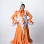 Traje de flamenca de la Colección 2026 en color naranja con lunares, diseño tradicional con volantes rematados en blanco y mantoncillo de flecos largos, elegante y luminoso.