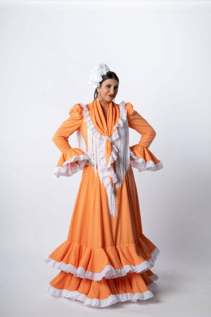 Traje de flamenca de la Colección 2026 en color naranja con lunares, diseño tradicional con volantes rematados en blanco y mantoncillo de flecos largos, elegante y luminoso.